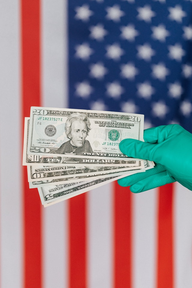 medical gloved hand holding American bills in front of an american flag