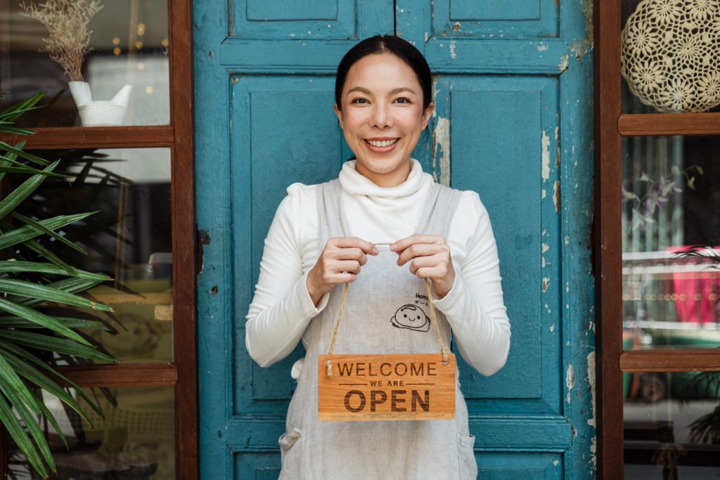 woman in pinafore in front of a blue door holding a sign that says welcome we are open